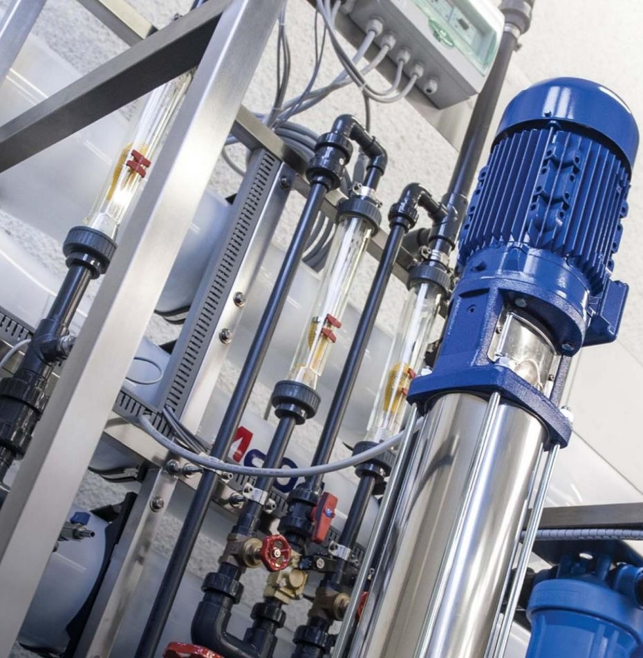 Kompleksowa instalacja przemysłowa z rurami, zaworami i niebieskim silnikiem elektrycznym, widoczne szklane cylindry z pomiarami poziomu cieczy, metalowa rama konstrukcyjna.