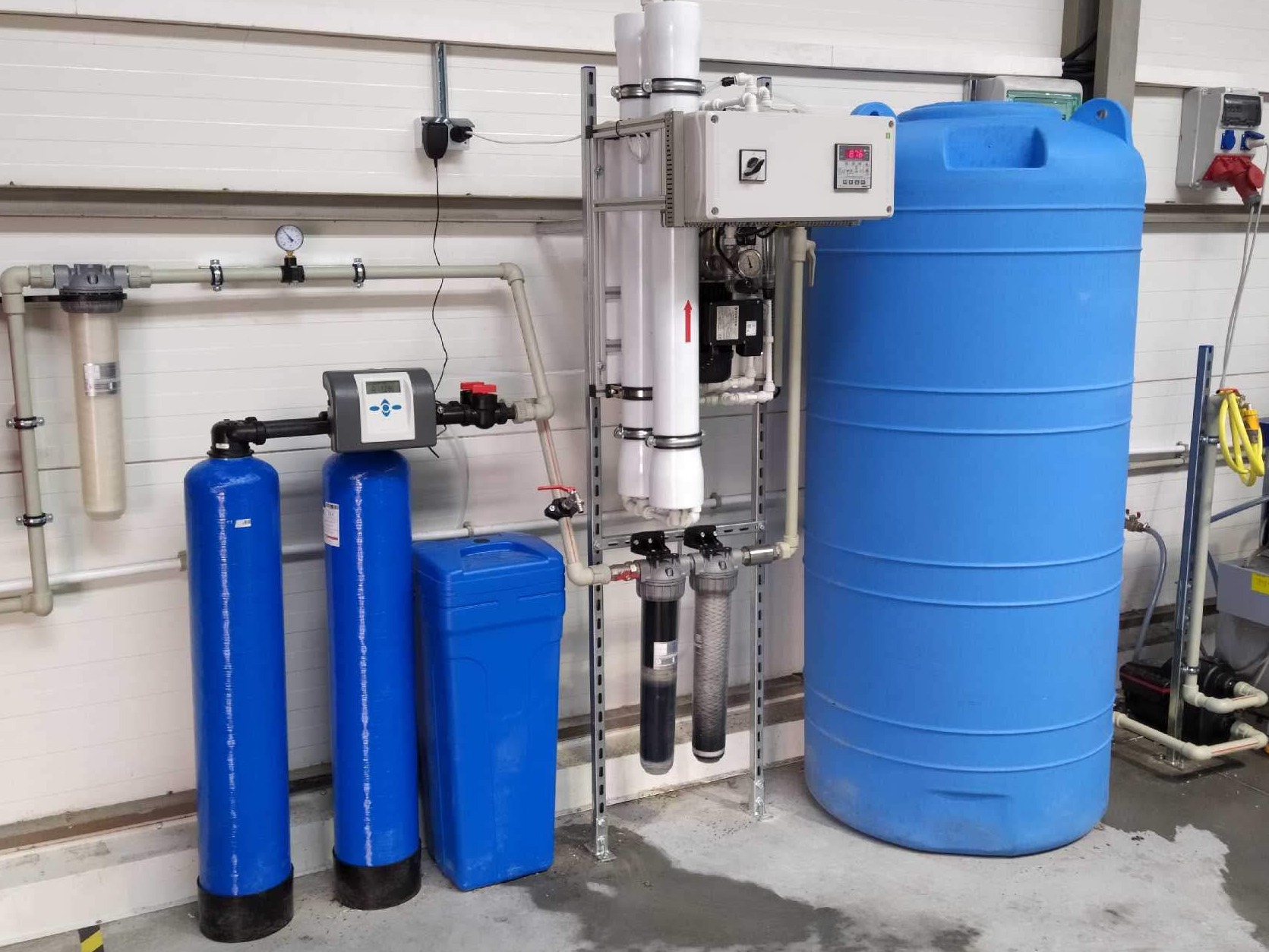 Kompletna instalacja filtracji wody z dużym niebieskim zbiornikiem, rurami i panelami sterującymi zamontowana w pomieszczeniu z płytami warstwowymi.