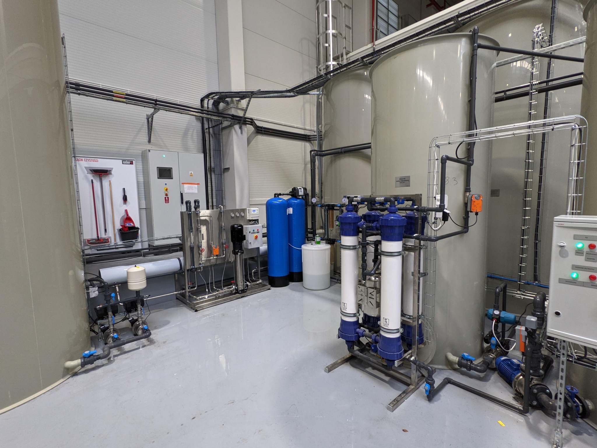 Instalacja przemysłowa z dużymi zbiornikami, filtrami i panelami sterowania w hali oświetlonej światłem dziennym. Widoczne rury, kable i elementy armatury. Na ścianie tablica z narzędziami...