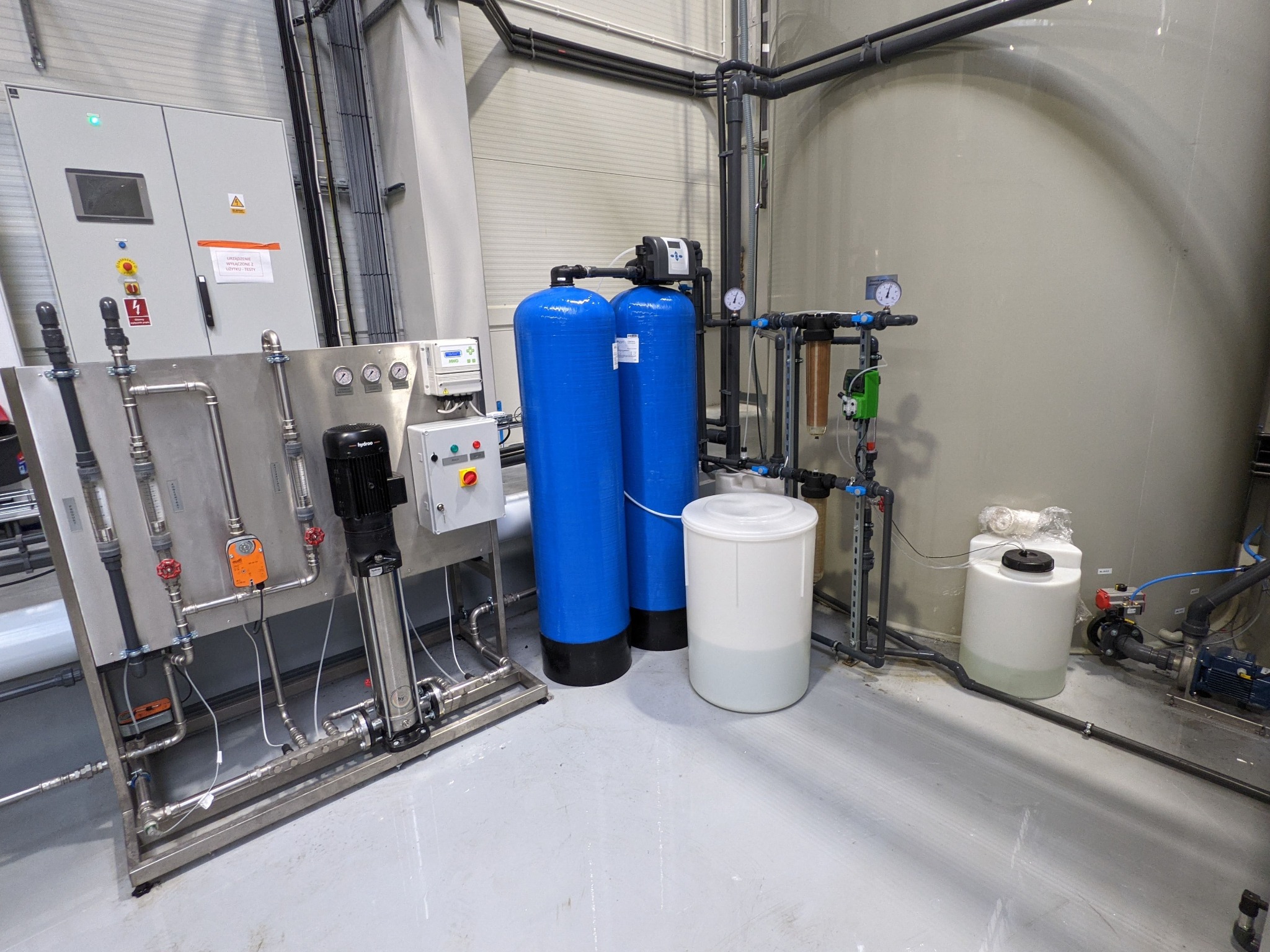 Kompletna instalacja uzdatniania wody z dwoma niebieskimi zbiornikami ciśnieniowymi, pompą i zbiornikami na wodę, umieszczona w hali z szarym betonowym podłożem i widocznymi rurami pod sufitem.