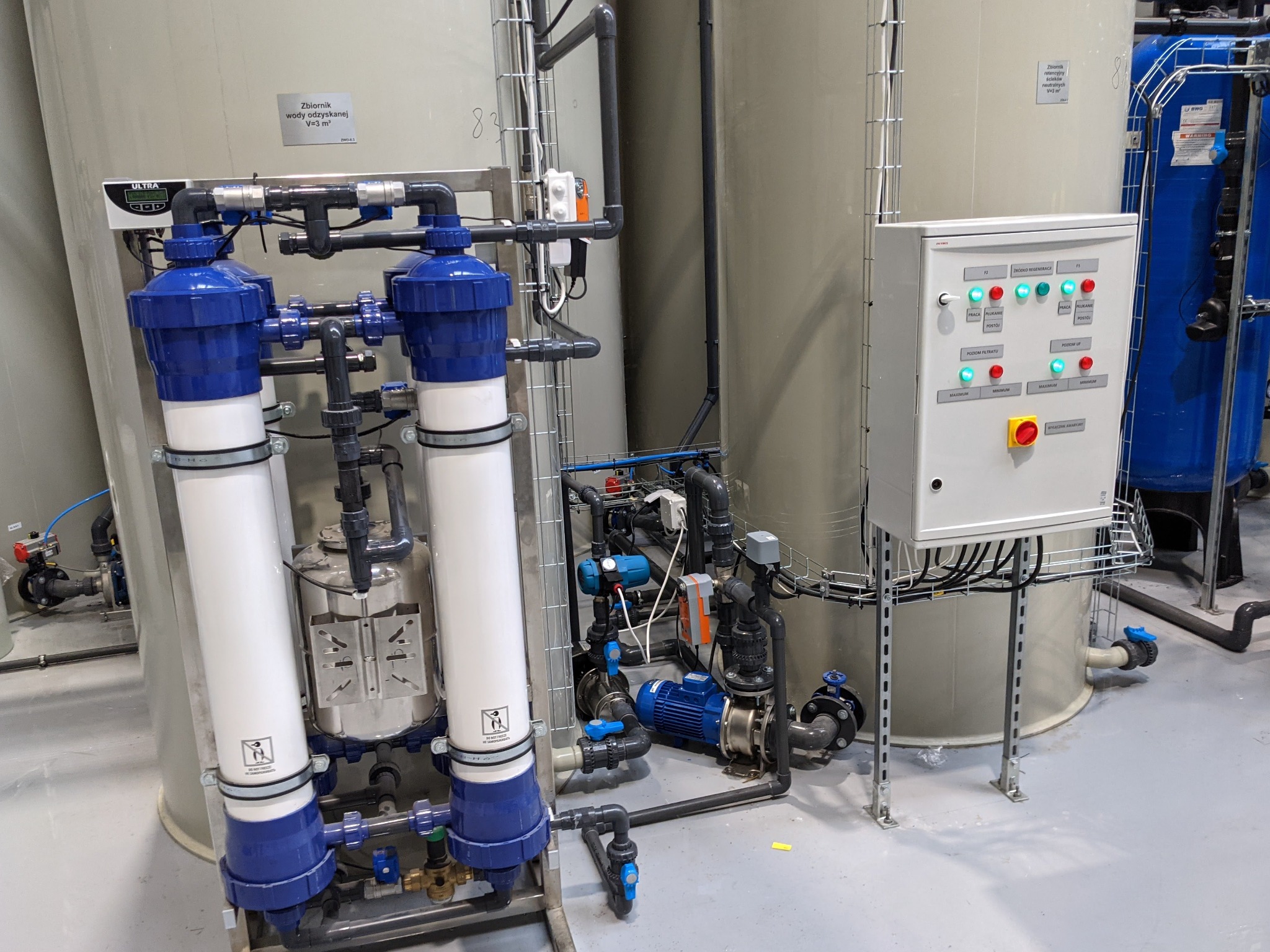Kompleksowa instalacja przemysłowa z dwoma pionowymi filtrami z niebieskimi elementami, zbiornikami i szafą sterowniczą z przyciskami i kontrolkami, widok z przodu.