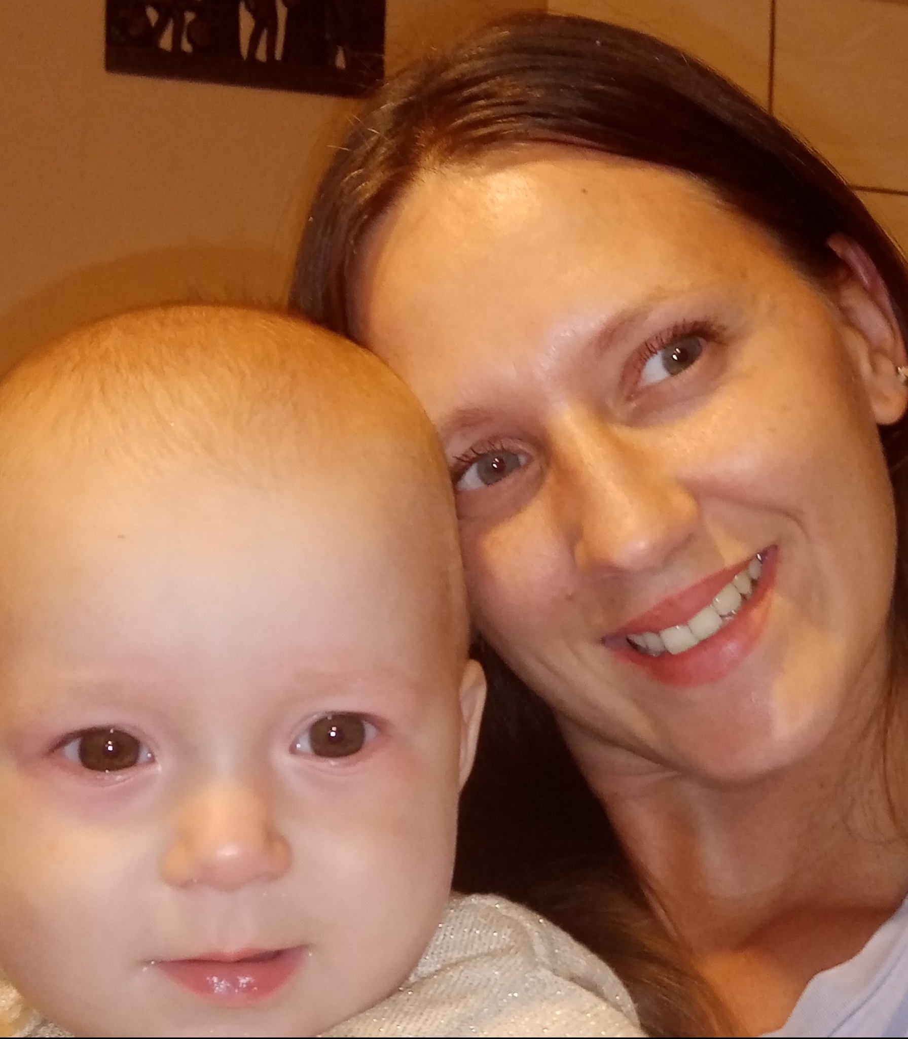 Portret uśmiechniętej kobiety z ciemnymi włosami obejmującej małe dziecko z jasnymi włosami, na rozmytym tle ściany w ciepłych odcieniach.