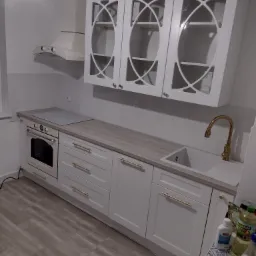 Biała zabudowa kuchenna z frezowanymi frontami szafek, złotymi uchwytami i baterią, widok na płytę grzewczą i okap.