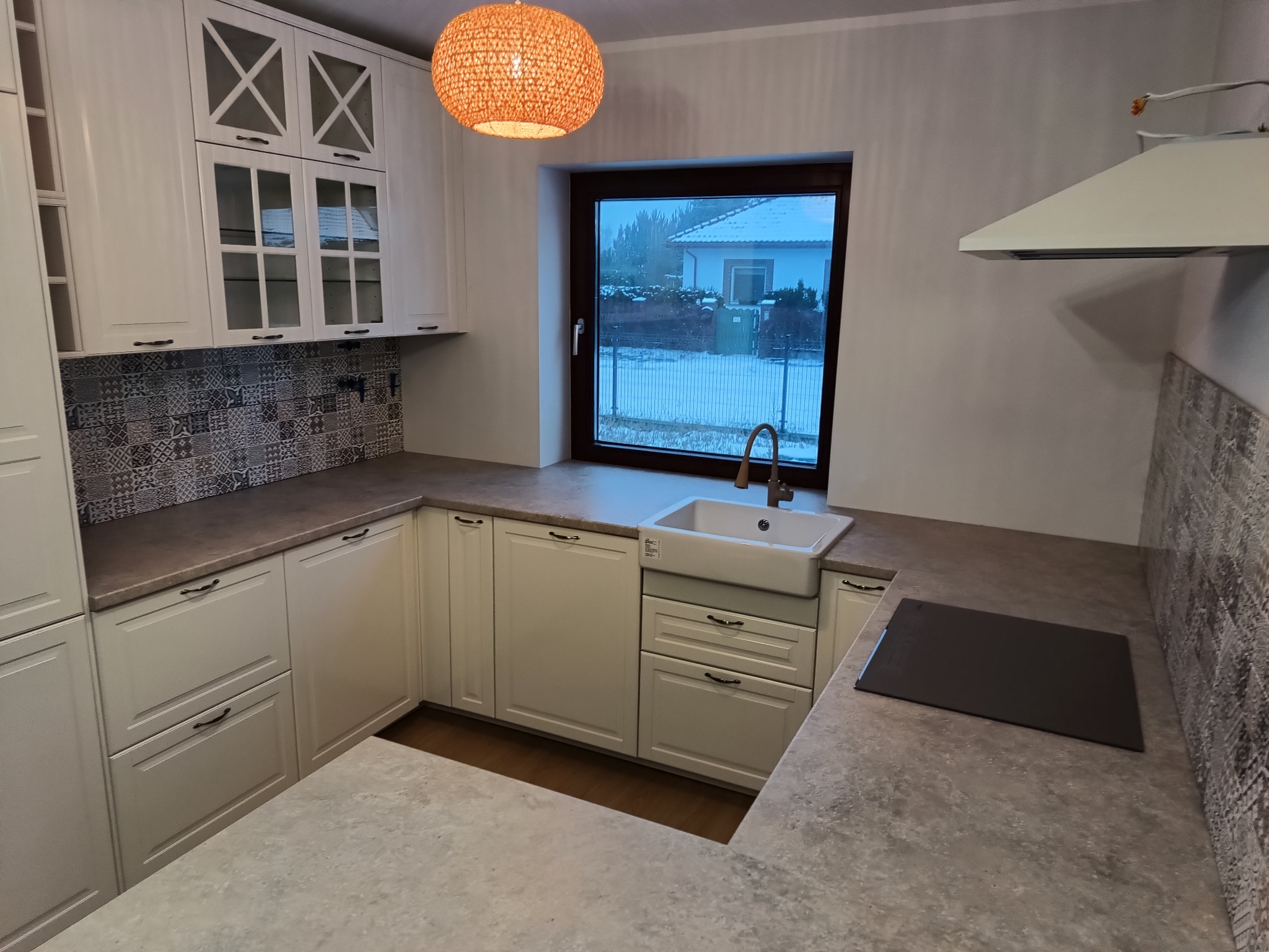 Jasna kuchnia z białymi szafkami, wzorzystą mozaiką nad blatem i widokiem na zimowy krajobraz za oknem, oświetlona ozdobną lampą z plecionki.