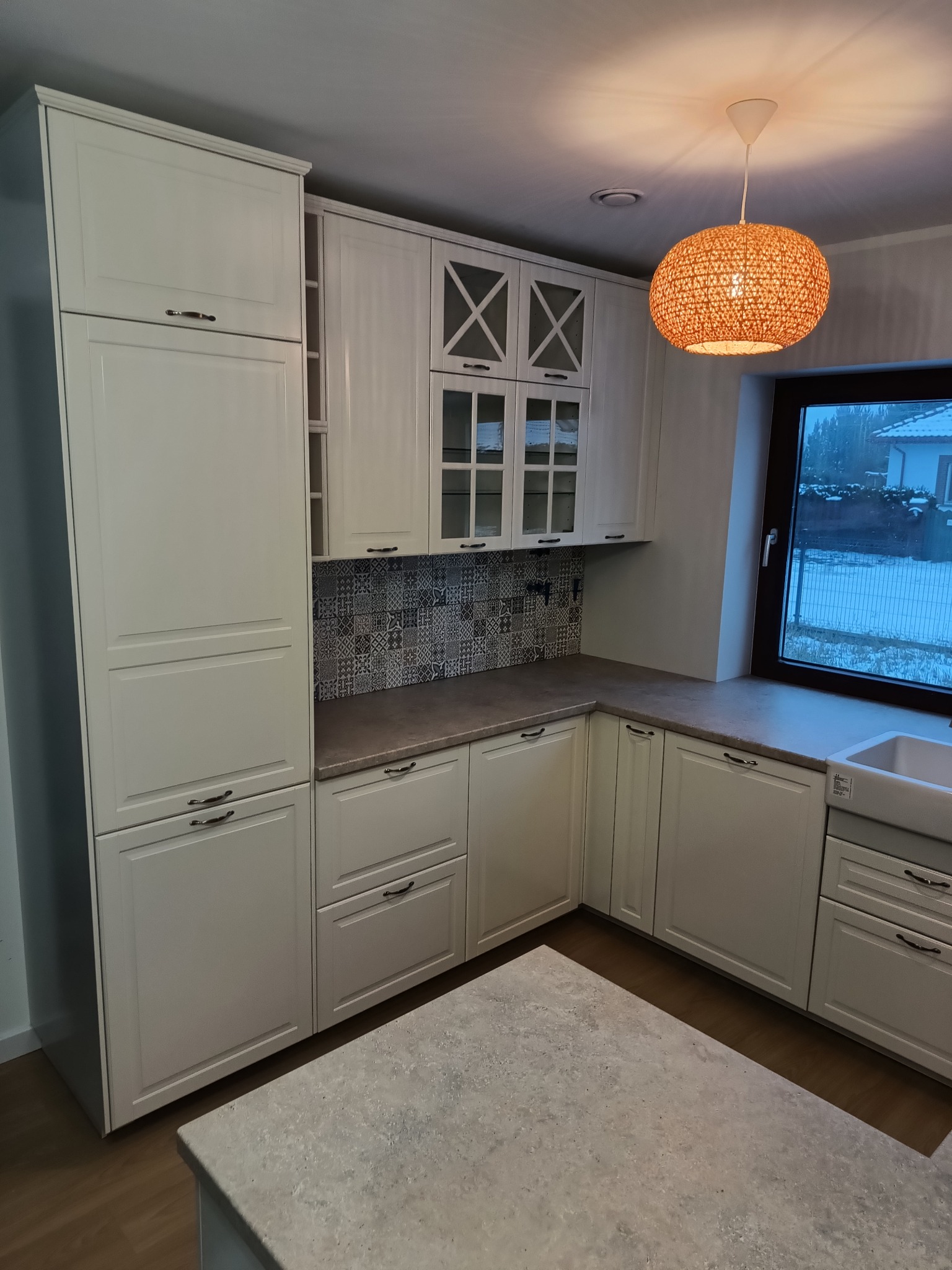 Jasna, nowoczesna kuchnia z białymi szafkami, wzorzystą mozaiką na ścianie nad blatem i okrągłą, plecioną lampą wiszącą. Za oknem widoczny zimowy krajobraz.