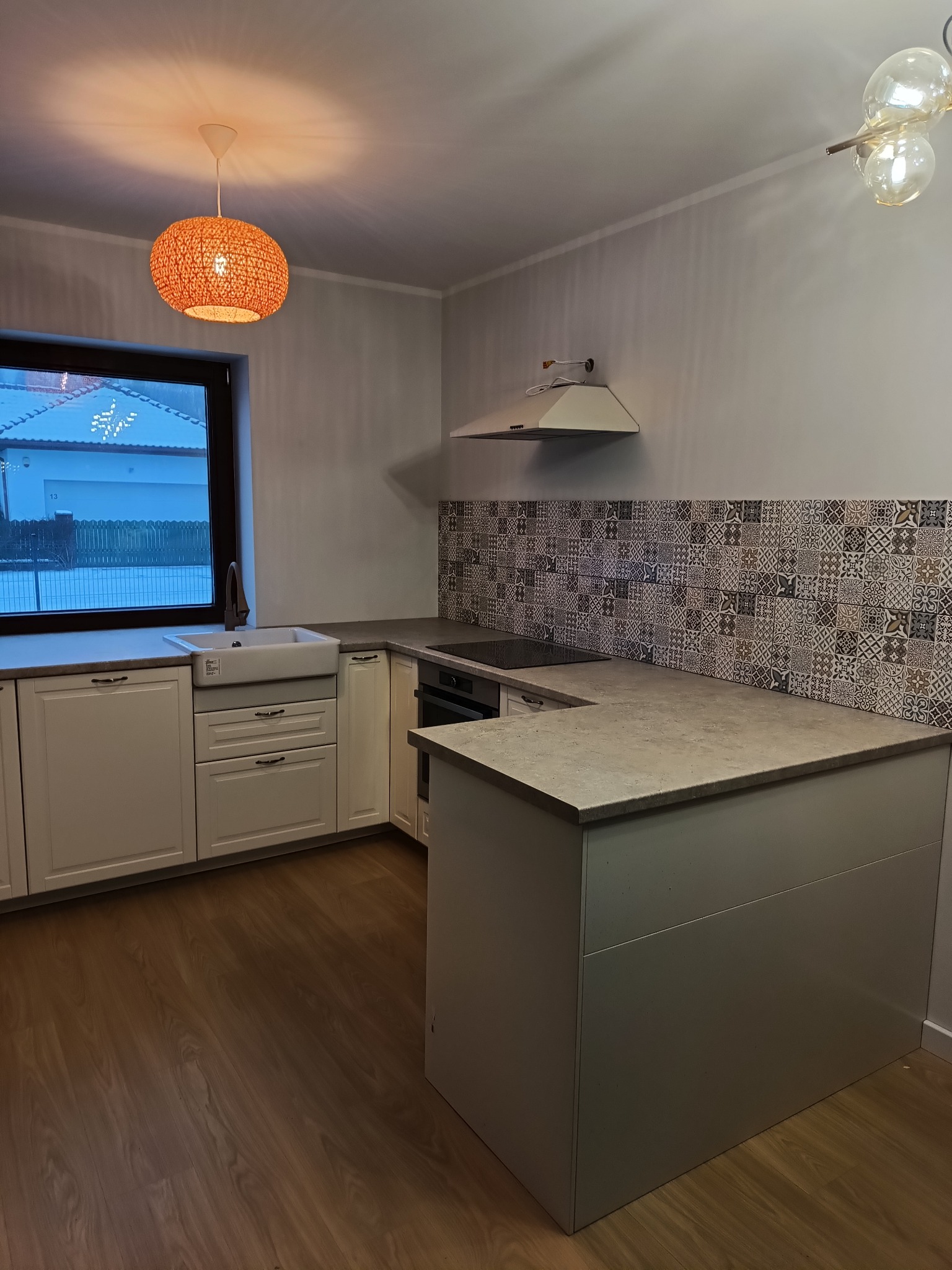 Jasna kuchnia z drewnianą podłogą, białymi szafkami z szarym blatem, wzorzystą ścianą nad blatem i dwoma różnymi lampami sufitowymi.