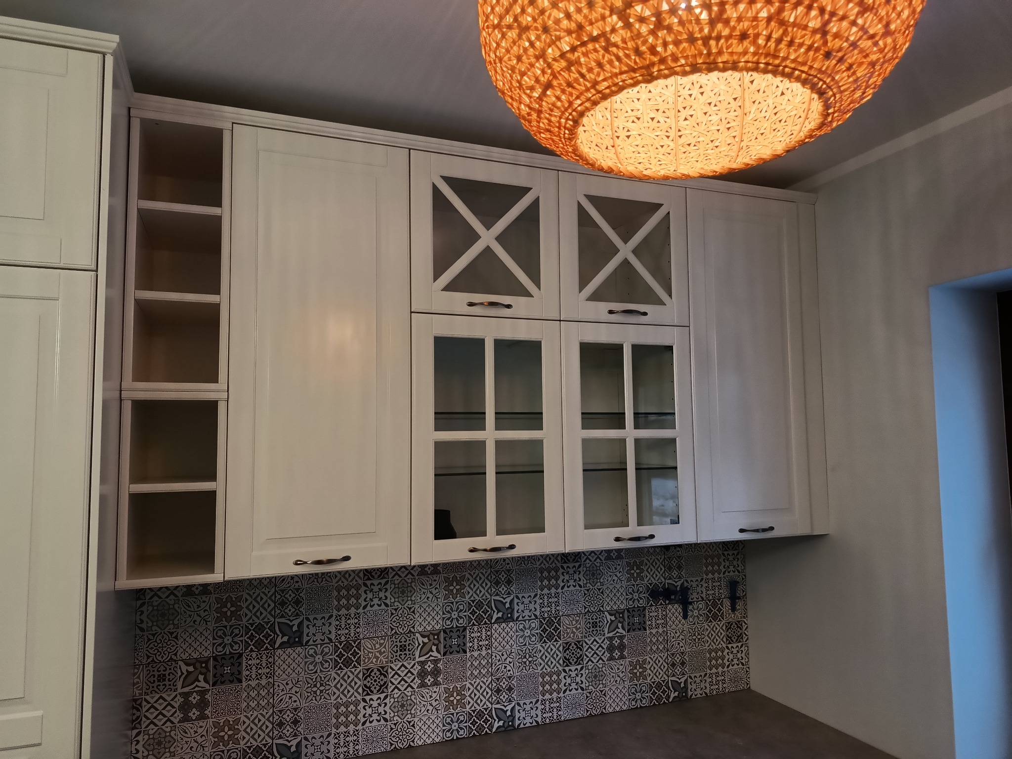 Białe szafki kuchenne z przeszklonymi frontami i dekoracyjnymi krzyżami, podświetlone okrągłą lampą z plecionki, nad blatem z mozaiką płytek.