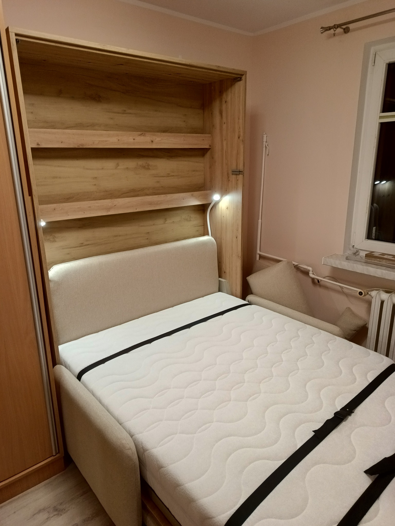 Rozkładane łóżko w szafie z oświetleniem punktowym, zabezpieczone pasami transportowymi, widoczne półki wewnątrz szafy.