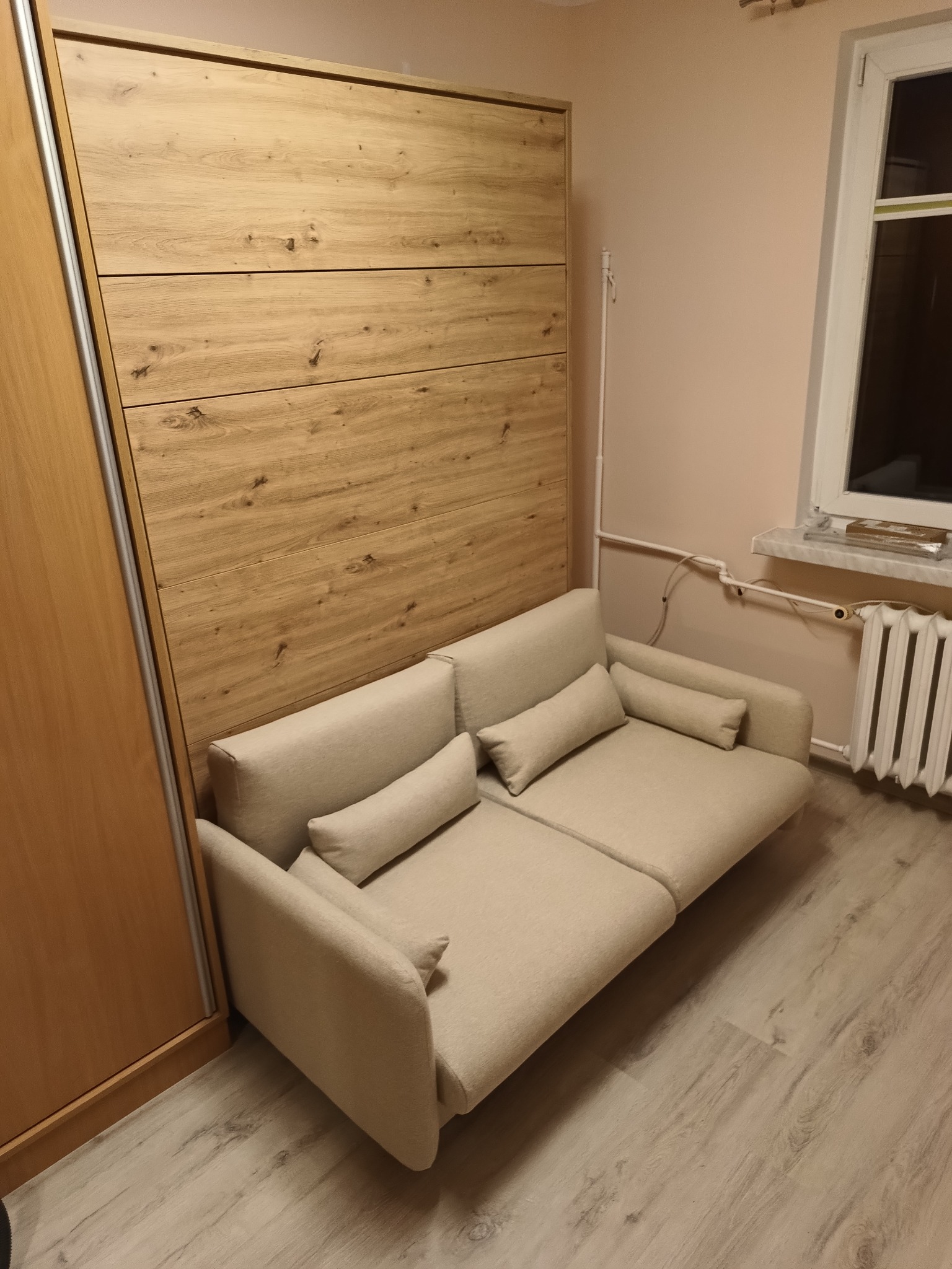 Sofa rozkładana zintegrowana z szafą wnękową o drewnianym froncie, widoczna w pokoju z grzejnikiem pod oknem.