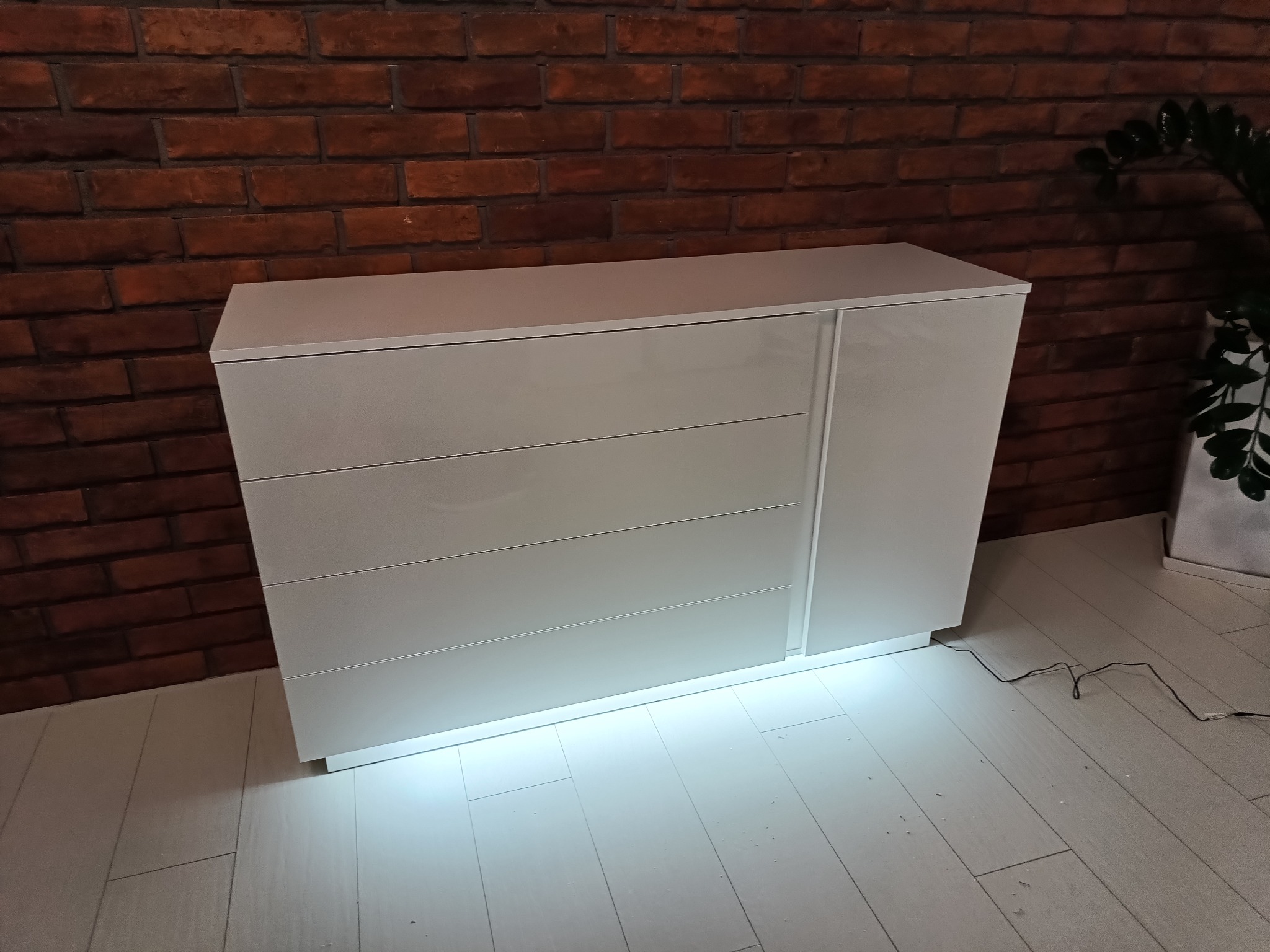 Biała komoda z trzema szufladami i bocznymi drzwiczkami na tle ceglanej ściany i jasnej podłogi, oświetlona od dołu.