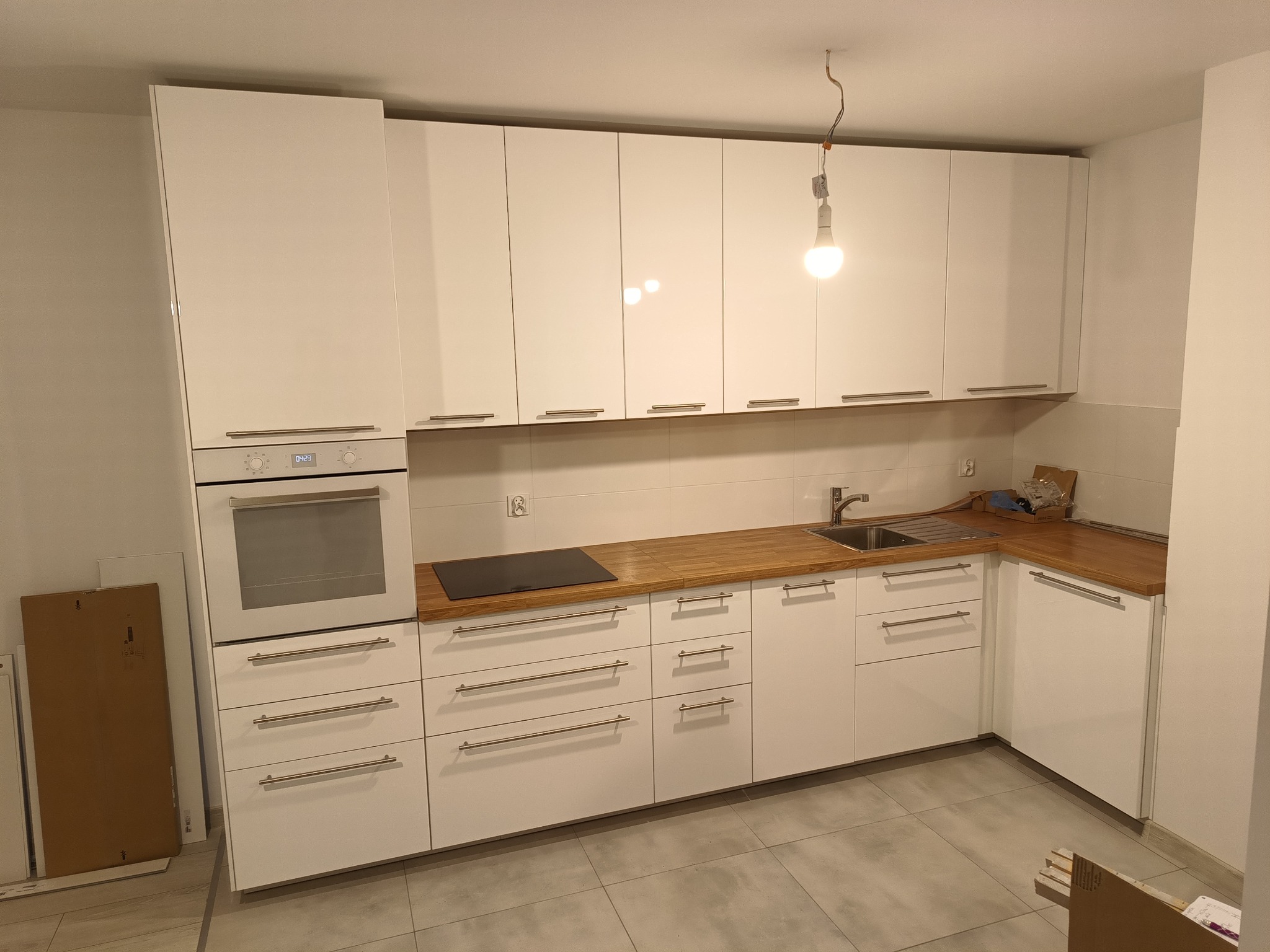 Nowoczesna, biała zabudowa kuchenna z drewnianym blatem i niedokończonym montażem oświetlenia wiszącego na kablu.
