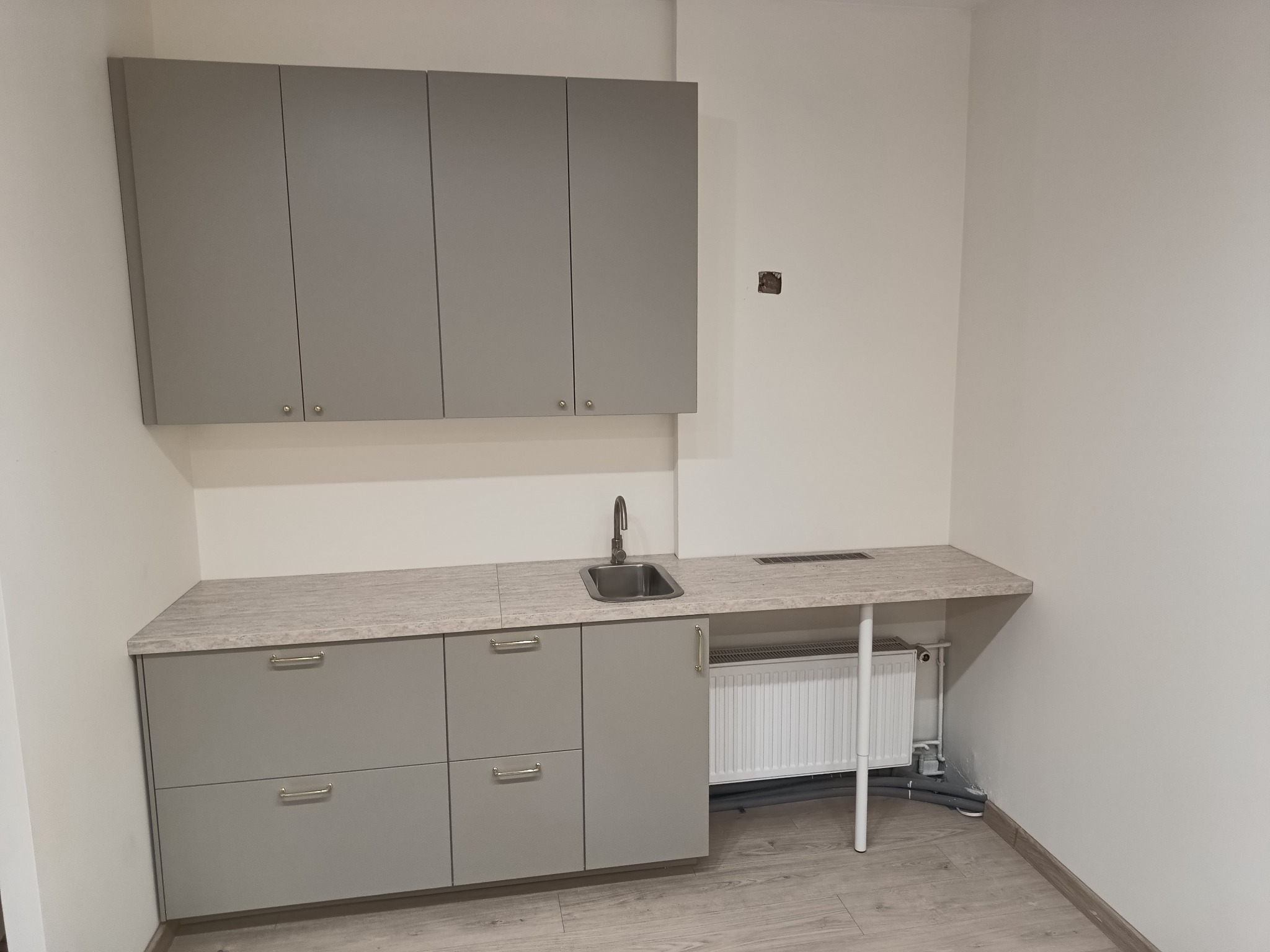 Szare szafki kuchenne wiszące i stojące z blatem w jasnym odcieniu drewna, widoczny zlew i grzejnik pod blatem, w tle biała ściana z widocznym gniazdkiem elektrycznym.