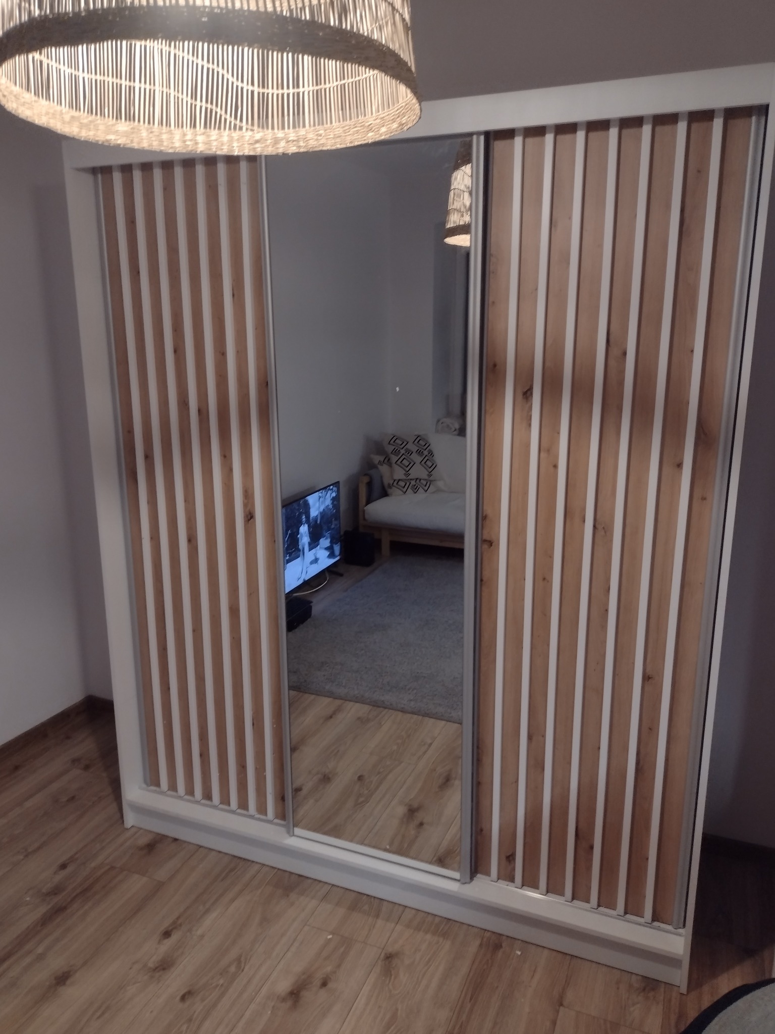 Nowoczesna szafa przesuwna z lustrem i drewnianymi lamelami, odbicie wnętrza pokoju z sofą i telewizorem.