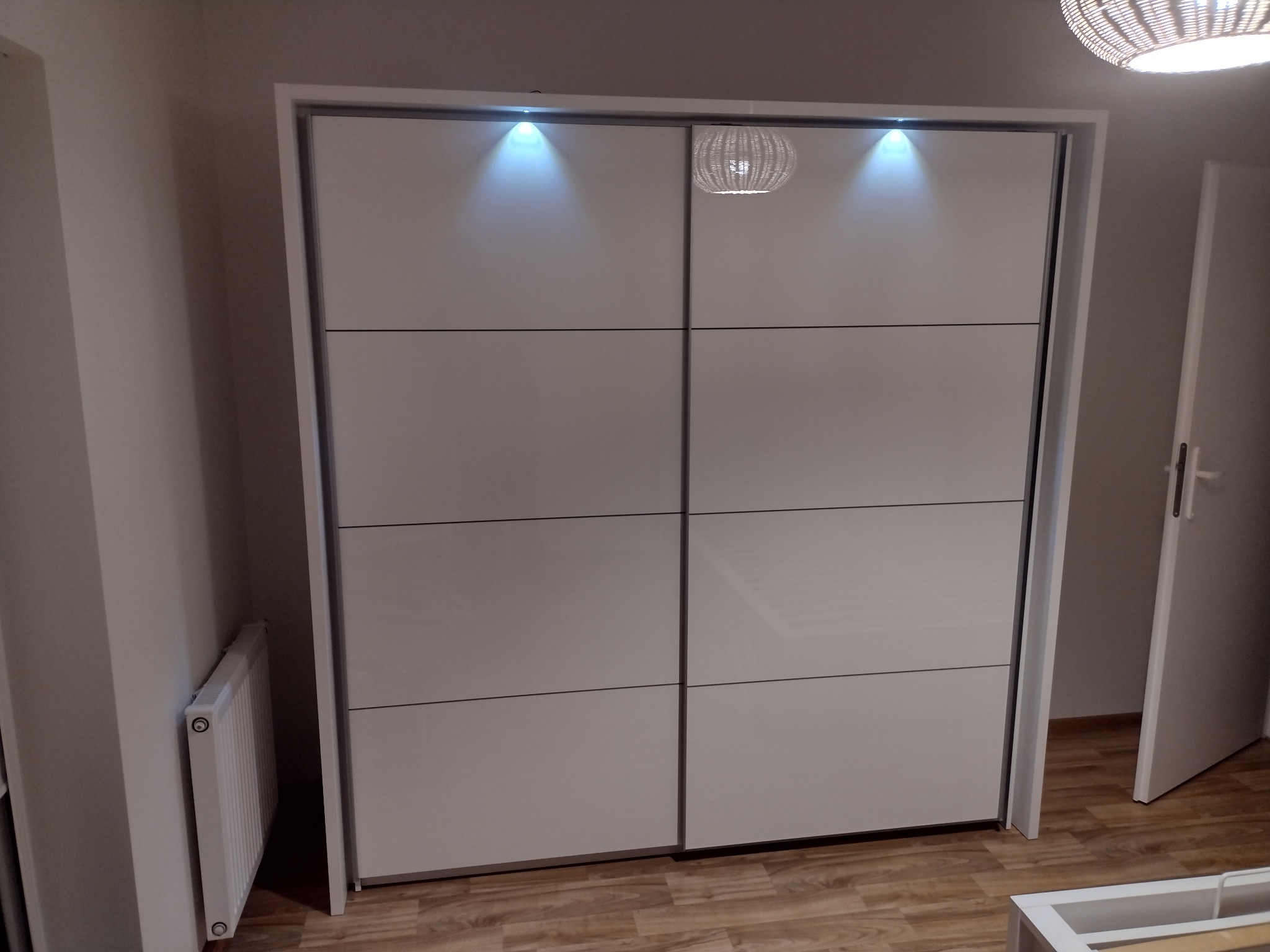 Biała szafa przesuwna z oświetleniem LED w górnej części, widoczna lampa wisząca, grzejnik i fragment otwartych drzwi.