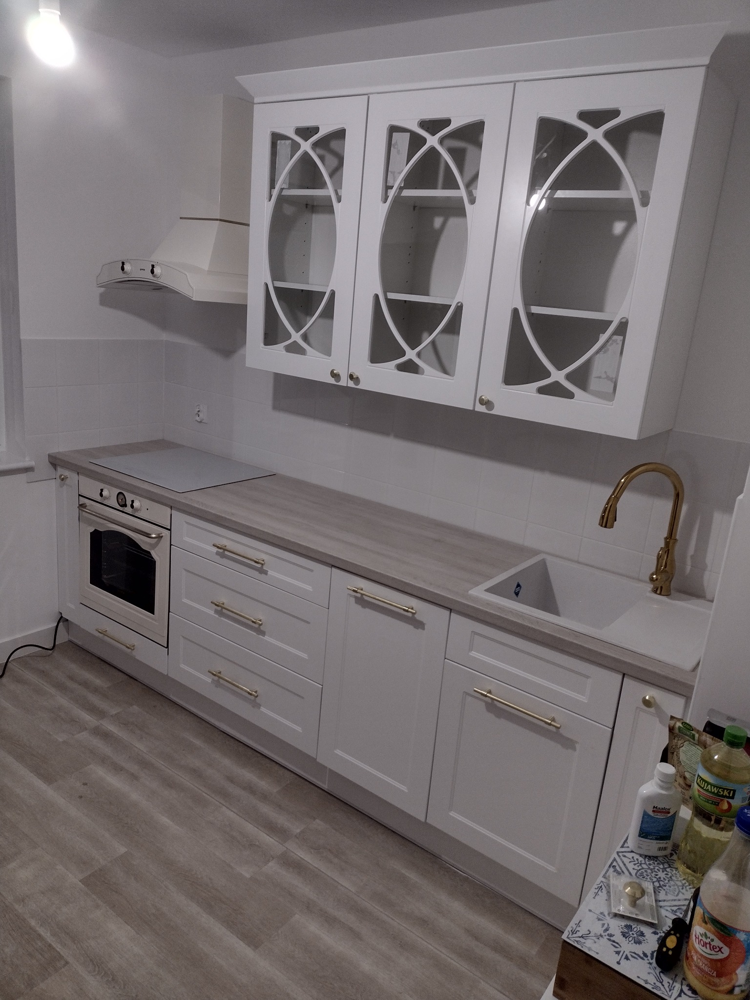 Biała zabudowa kuchenna z frezowanymi frontami szafek, złotymi uchwytami i baterią, widok na płytę grzewczą i okap.