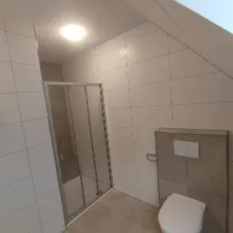 Wykończona łazienka na poddaszu z kabiną prysznicową, wiszącą toaletą z zabudową w odcieniach szarości i białymi płytkami na ścianach.