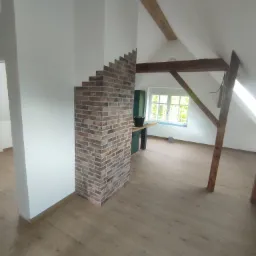 Poddasze po remoncie: wykończona ściana z cegły klinkierowej, drewniana podłoga, widoczne elementy konstrukcji dachu i okno.
