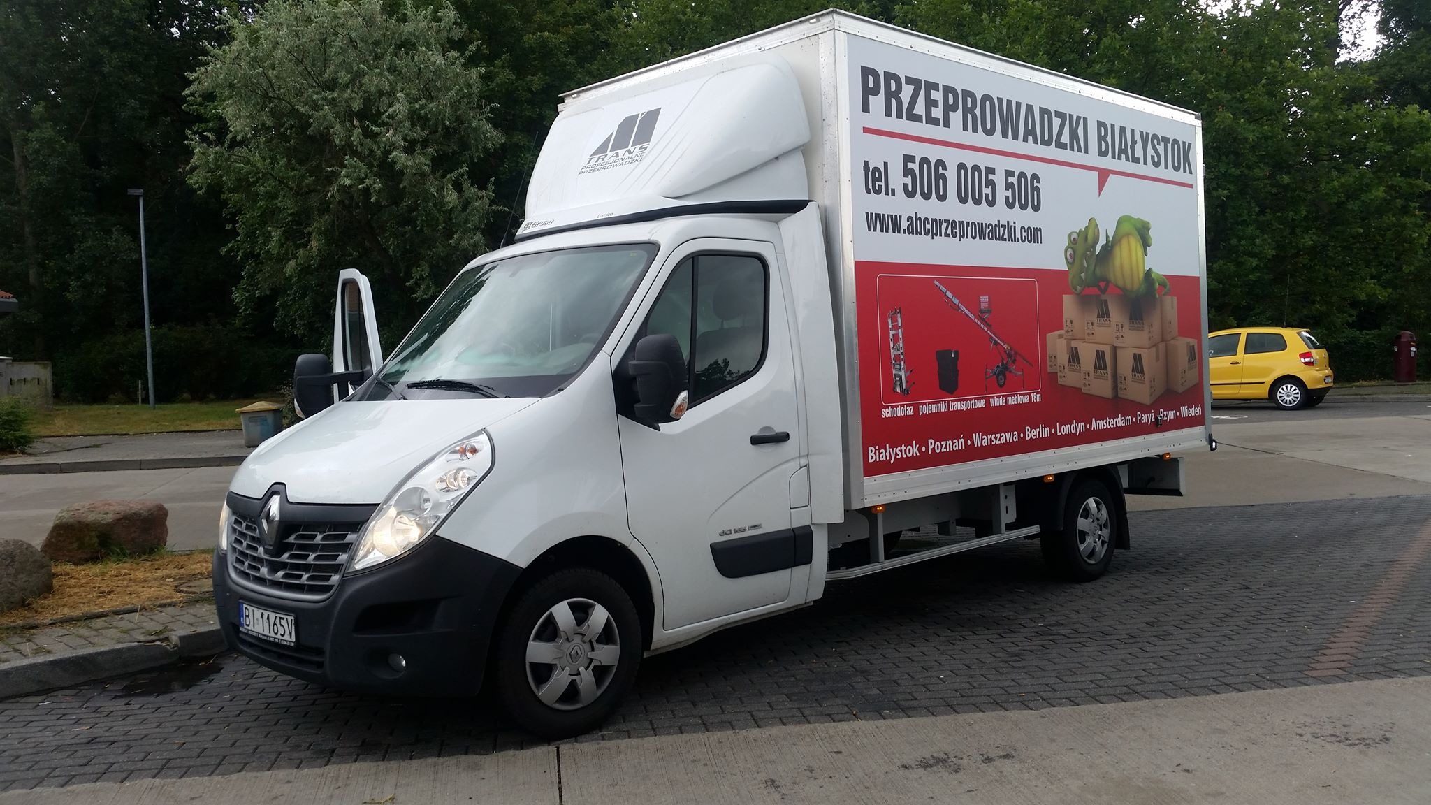 Biały samochód dostawczy Renault z otwartymi drzwiami kierowcy i reklamą firmy przeprowadzkowej z Białegostoku, na której widoczny jest schodołaz, pojemniki transportowe i winda meblowa, w tle...