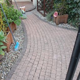 Daniel Dubis - Zakrzywiony chodnik z kostki brukowej w ogrodzie, obok roślin w donicach i worka ziemi. Widoczna szczotka do zamiatania. Ujęcie z poziomu gruntu.