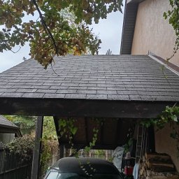 Daniel Dubis - Szary dach pokryty papą bitumiczną, widoczny z dołu, z fragmentem zadaszenia garażowego i samochodem. W tle drzewa i fragment budynku.