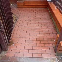 Daniel Dubis - Czerwona kostka brukowa po umyciu, mokra nawierzchnia, widoczne ślady wody i piany, brama z metalowymi prętami po lewej stronie kadru, schody z kostki w tle.