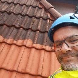 Daniel Dubis - Częściowo umyty dach z dachówki ceramicznej, widoczna różnica w kolorze, pracownik w kasku i odblaskowej kurtce, ujęcie z dołu.