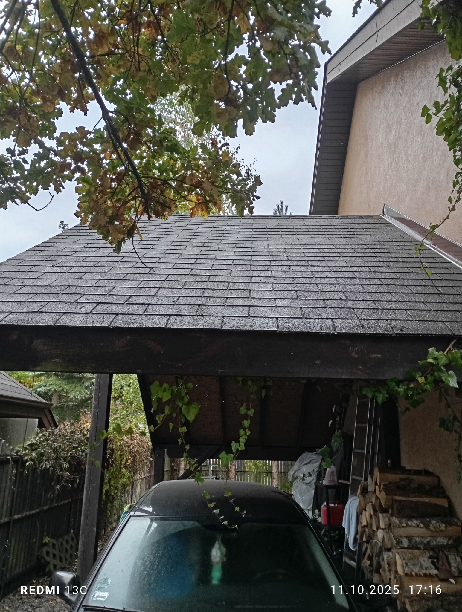 Szary dach pokryty papą bitumiczną, widoczny z dołu, z fragmentem zadaszenia garażowego i samochodem. W tle drzewa i fragment budynku.