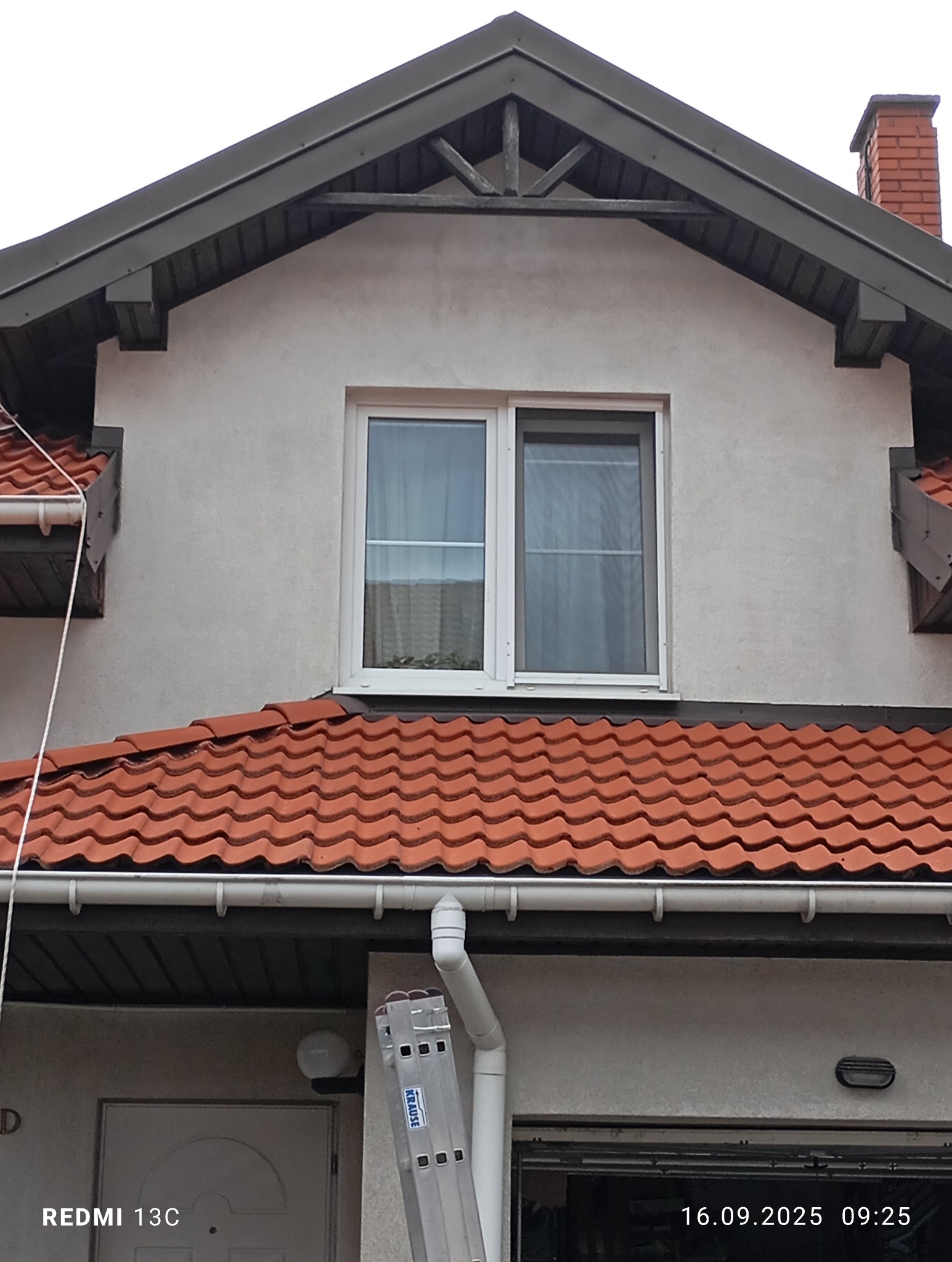 Dwukondygnacyjny dom z czerwonym dachem i białą elewacją. Widoczna drabina oparta o rynnę, sugerująca prace konserwacyjne lub czyszczenie.