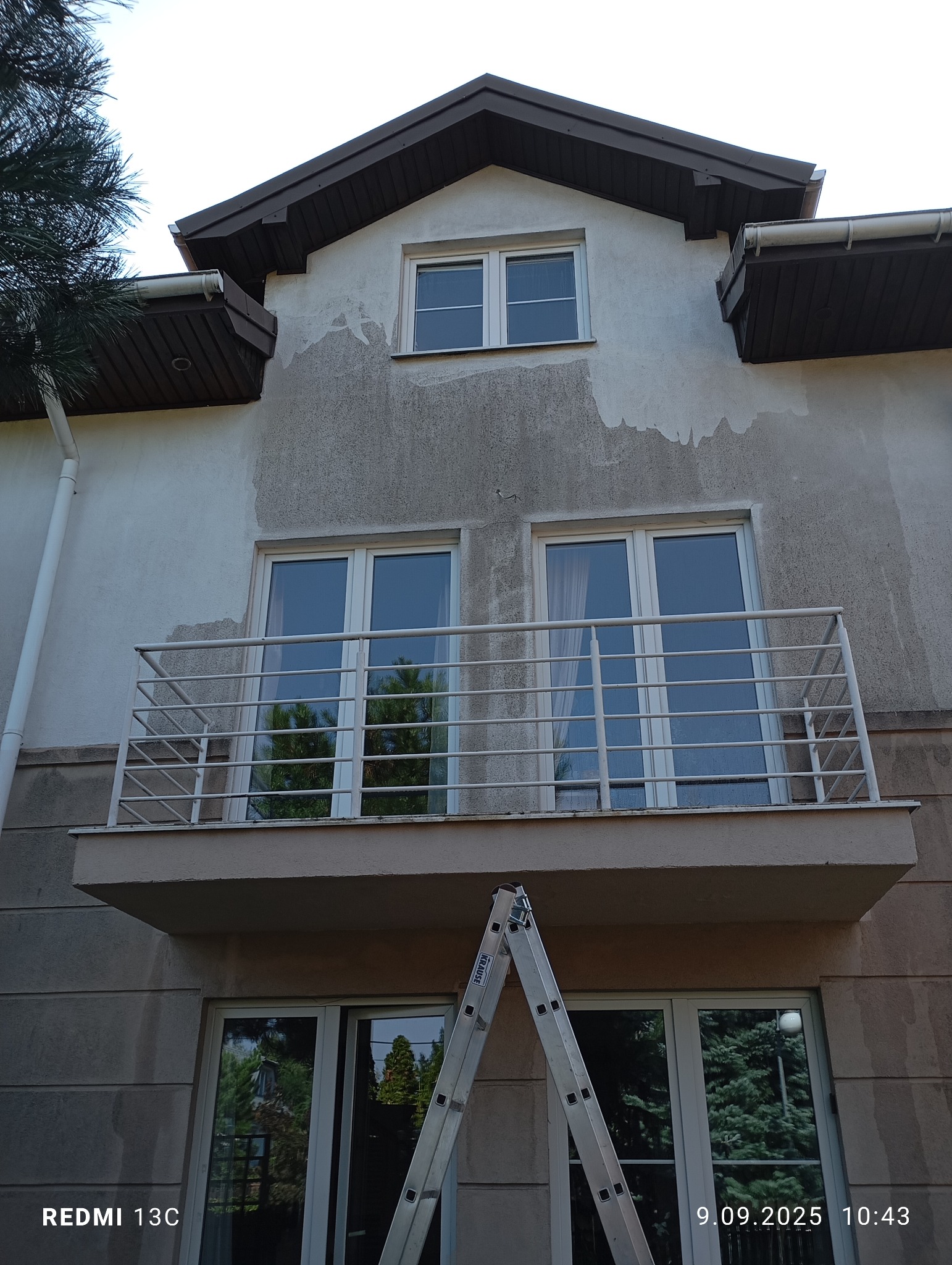 Dwupiętrowy dom z balkonem przed czyszczeniem elewacji. Widoczne zabrudzenia i drabina oparta o ścianę. Ujęcie z dołu, podkreślające wysokość budynku.