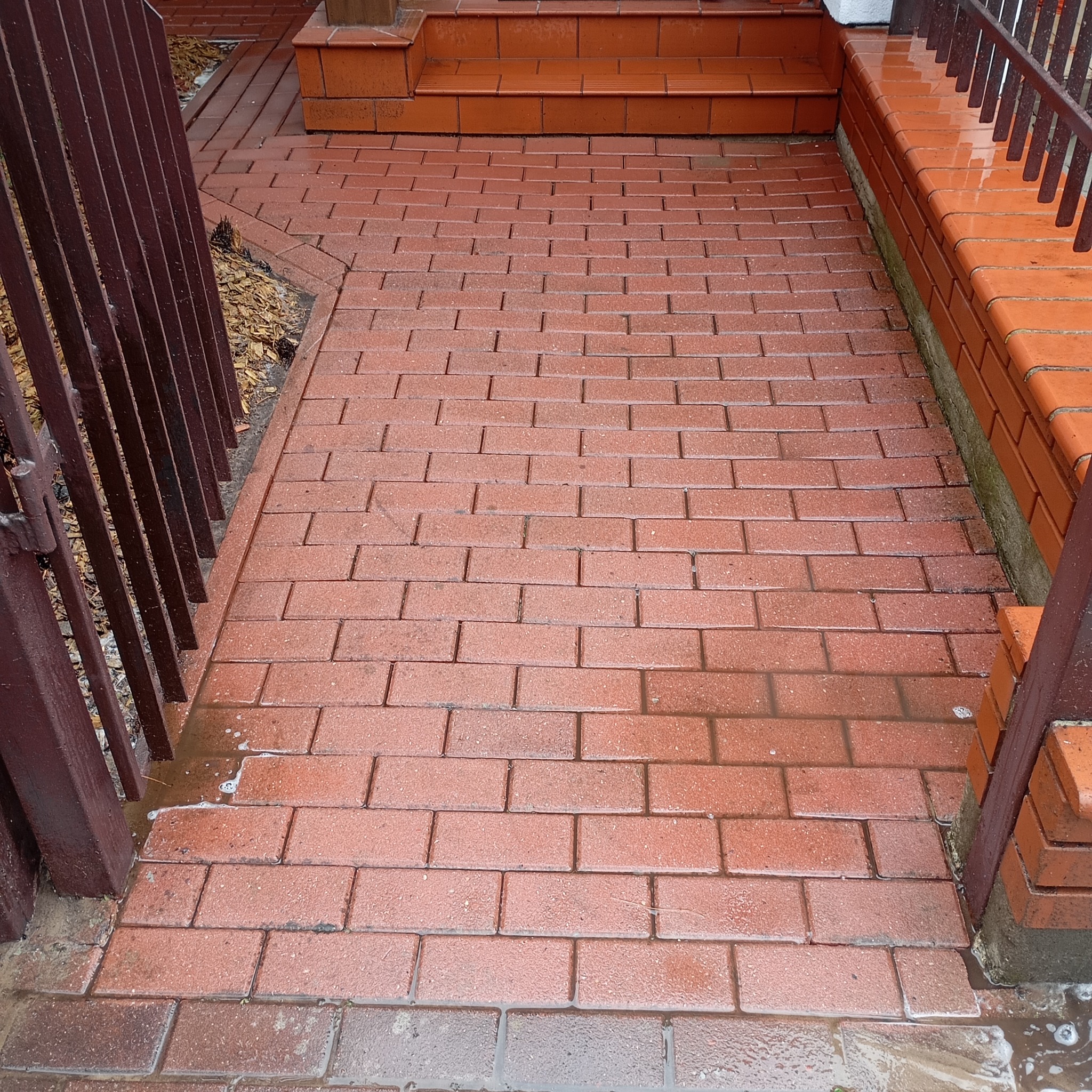 Czerwona kostka brukowa po umyciu, mokra nawierzchnia, widoczne ślady wody i piany, brama z metalowymi prętami po lewej stronie kadru, schody z kostki w tle.