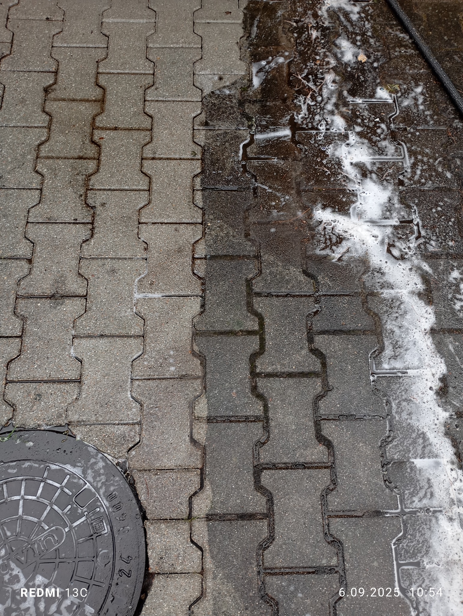 Mycie kostki brukowej: widoczna różnica między czystą, szarą kostką a mokrą, brudną. Część kostki pokryta pianą, w rogu studzienka kanalizacyjna.