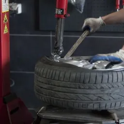 Demontaż opony z felgi przy użyciu montażownicy w warsztacie; widoczne ręce w rękawicach, opona, felga, montażownica i narzędzia.