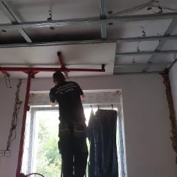 Pracownik montuje płytę gipsowo-kartonową na metalowym stelażu sufitowym za pomocą podnośnika, widoczne ślady uszkodzeń na ścianach i okno z widokiem na zieleń.