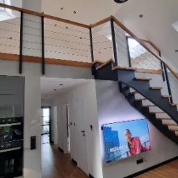 Nowoczesne schody z czarną konstrukcją, drewnianymi stopniami i balustradą z cienkich, poziomych linek, prowadzące na antresolę w jasnym wnętrzu z oknami dachowymi.