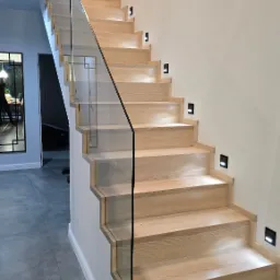 Nowoczesne schody drewniane z jasnego drewna z minimalistyczną balustradą ze szkła i czarnymi akcentami, oświetlone małymi lampkami LED w ścianie.