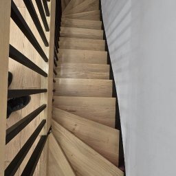 Stolarnia Rompa - Kątowe ujęcie dębowych schodów z czarnymi, metalowymi balustradami. Minimalistyczny design, jasne drewno i białe ściany. Widoczny fragment buta na pierwszym planie.