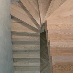 Stolarnia Rompa - Spiralne schody drewniane z jasnego drewna, widok z góry, betonowy sufit, parkiet w jodełkę, buty w dolnym rogu kadru. Nowoczesne wykończenie.
