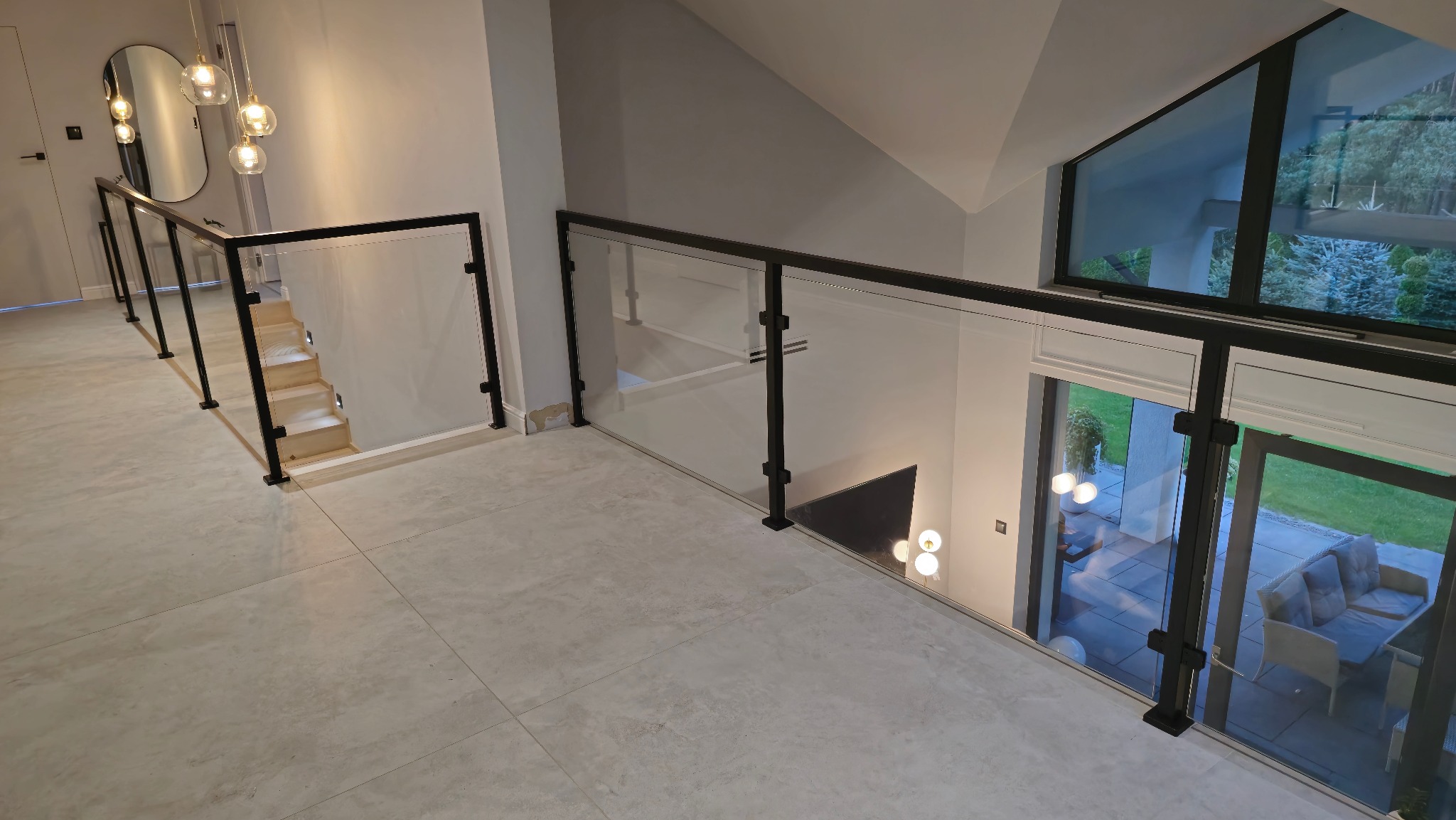 Nowoczesne wnętrze z balustradą szklaną z czarnymi słupkami, widok z góry na schody i salon z wyjściem na taras.