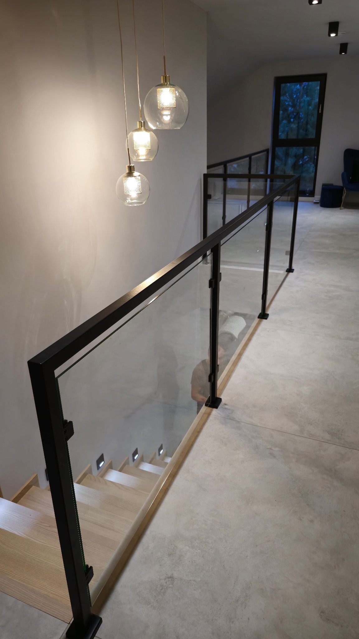 Nowoczesne schody z jasnego drewna z czarną, szklaną balustradą, oświetlone designerskimi lampami wiszącymi, widok z góry na betonową posadzkę.