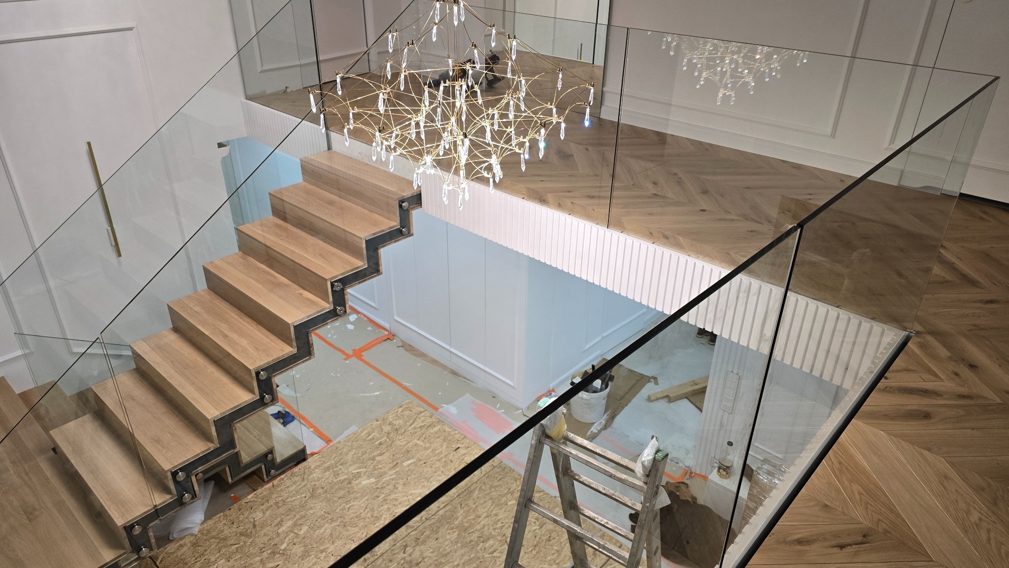 Nowoczesne schody z drewnianymi stopniami i szklanymi balustradami, widok z góry na klatkę schodową z dekoracyjnym żyrandolem i parkietem w jodełkę.