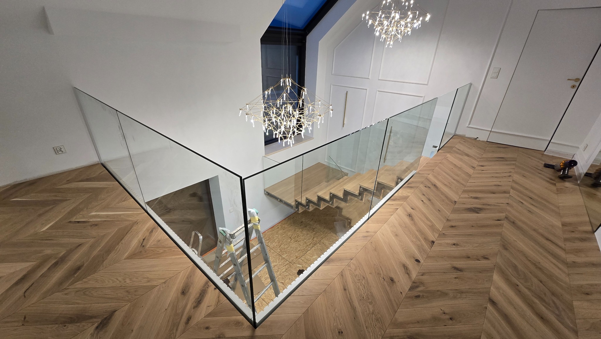 Nowoczesne schody z drewnianymi stopniami i szklaną balustradą, widok z góry na parkiet w jodełkę, drabina oparta o ścianę, żyrandol w tle.