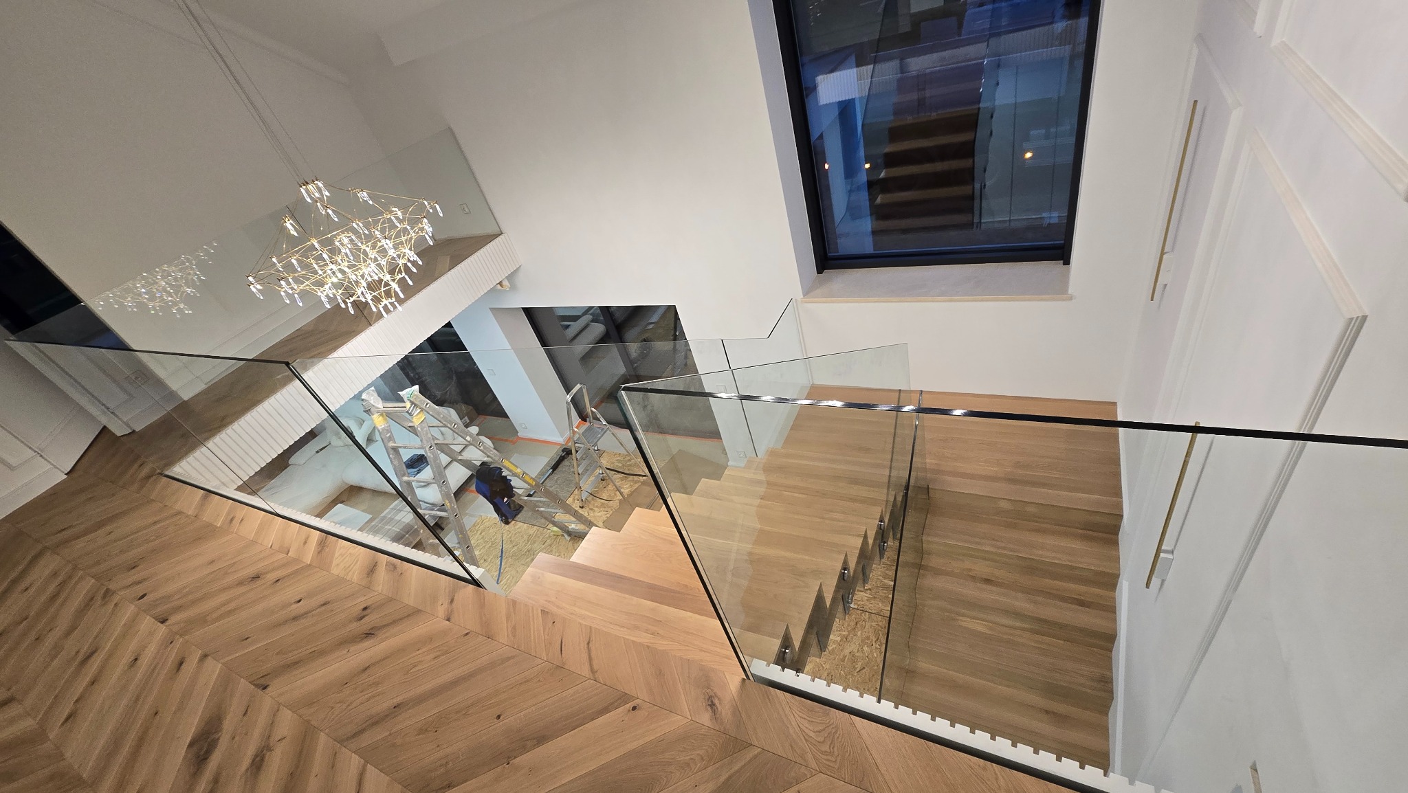 Nowoczesne schody z drewnianymi stopniami i szklaną balustradą, widok z góry, z elementami dekoracyjnymi i oknem w tle. Na podeście widoczna drabina.