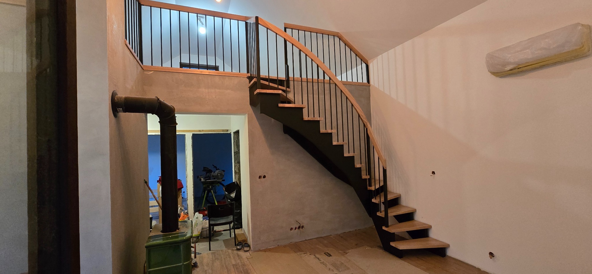 Spiralne schody z drewnianymi stopniami i czarną metalową balustradą w surowym wnętrzu z piecem i widoczną instalacją. Widok na antresolę.