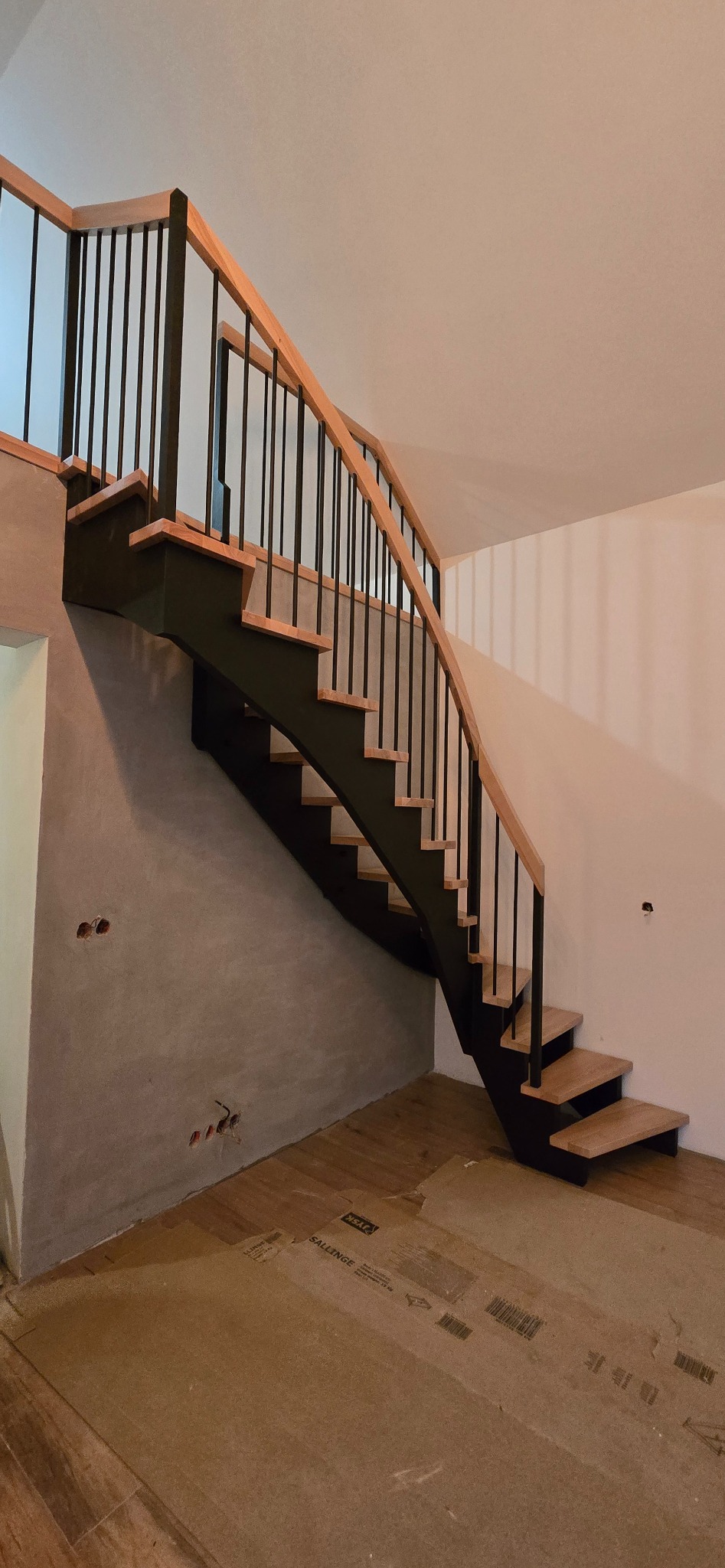 Nowoczesne schody z drewnianymi stopniami i czarną metalową balustradą, widok z dołu, surowy stan wykończenia wnętrza, zabezpieczona podłoga.