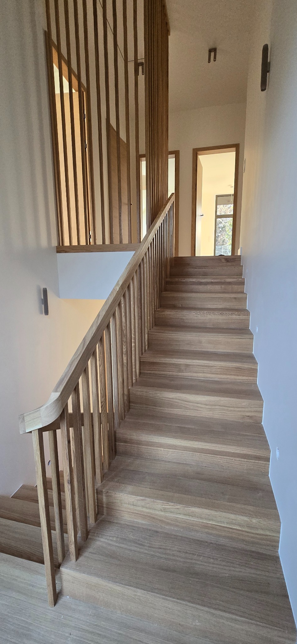 Jasne, drewniane schody z ażurową balustradą w nowoczesnym wnętrzu. Widok z dołu, schody prowadzą do jasnego pomieszczenia z widocznym oknem.