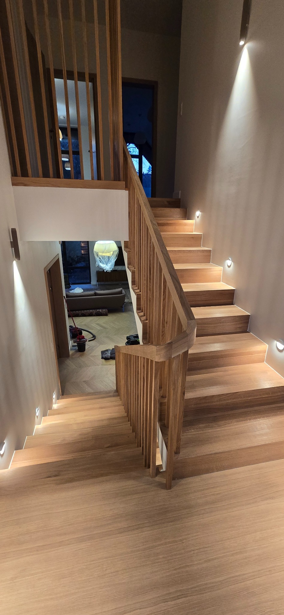 Jasne, drewniane schody z balustradą i oświetleniem punktowym wzdłuż ściany. Widok z góry na dół, z widocznym salonem w tle. Nowoczesny design.