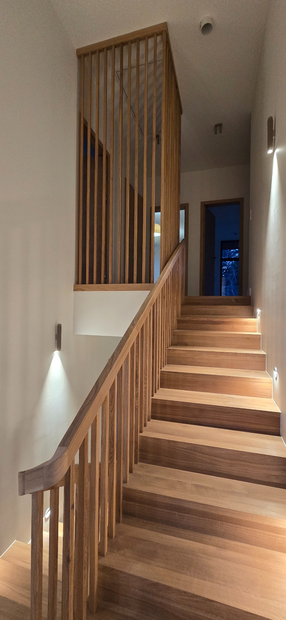 Jasne, drewniane schody z balustradą z pionowych szczebli, oświetlone punktowymi lampami ściennymi, prowadzące do otwartych drzwi w głębi korytarza.