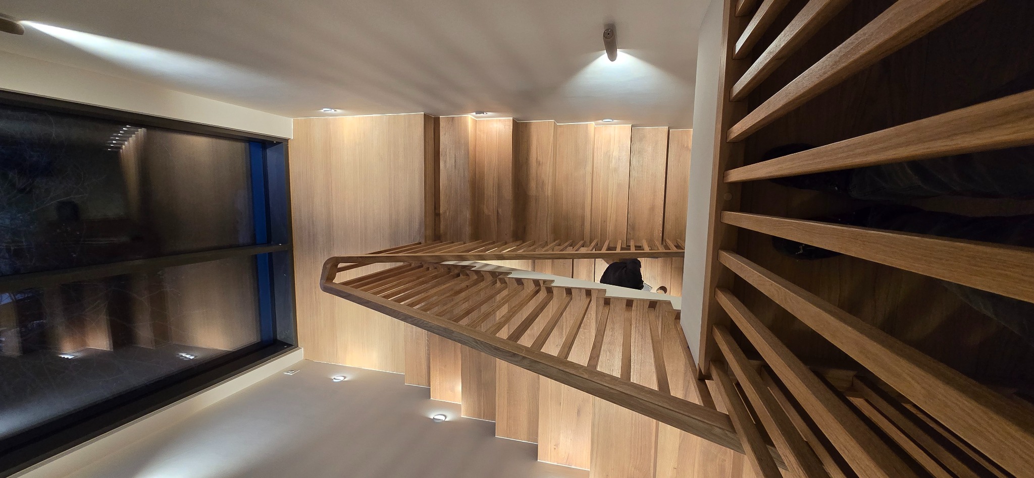 Nowoczesne schody drewniane z podświetleniem LED wzdłuż stopni i ściany, widok z góry, z elementami ażurowej balustrady i dużym oknem z boku.