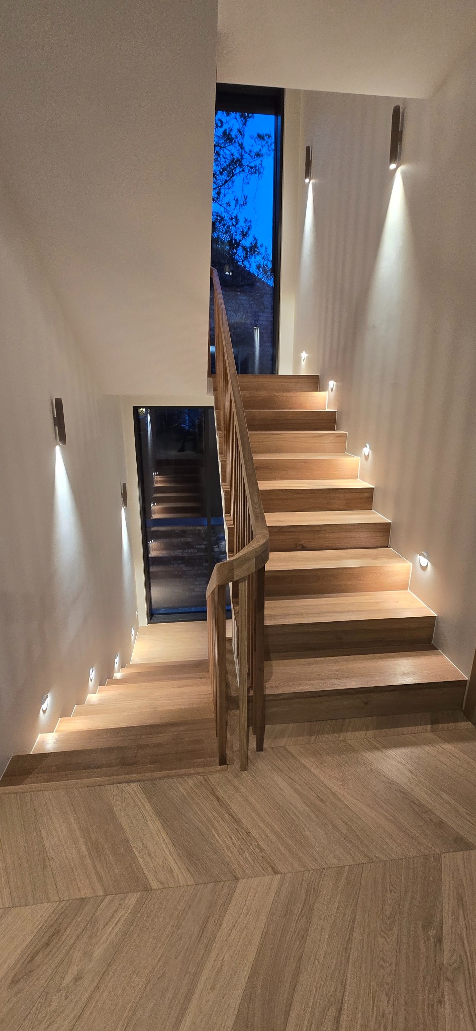 Nowoczesne, drewniane schody z podświetleniem LED wzdłuż stopni i na ścianie. Widok z góry na dół, parkiet ułożony w jodełkę. Okno z widokiem na nocne niebo.