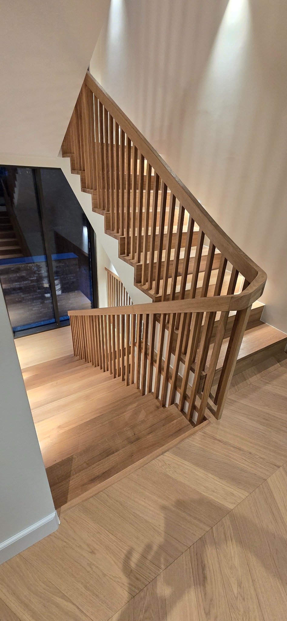 Jasne, drewniane schody z balustradą z pionowych listewek, widok z góry, parkiet w jodełkę na podłodze, nowoczesne oświetlenie punktowe.